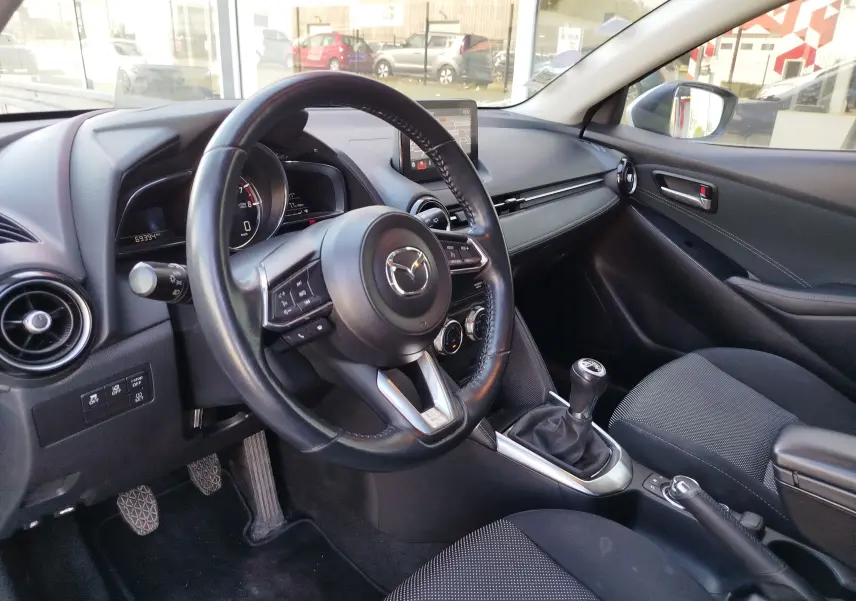 Vue intérieure côté conducteur de la Mazda2 2018, volant cuir, levier de vitesse manuel et tableau de bord moderne.