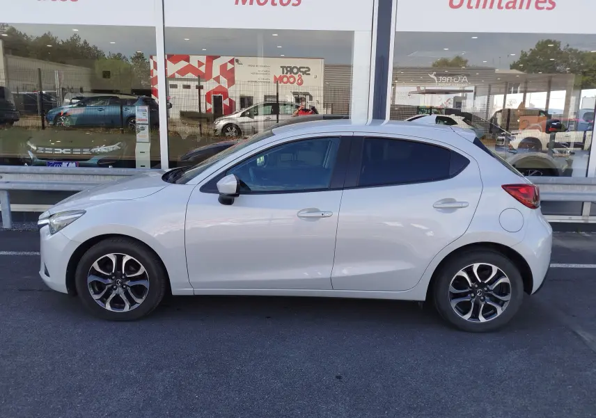 Vue de profil côté gauche d'une Mazda2 blanche nacrée avec jantes alliage bi-ton, stationnée devant une vitrine.