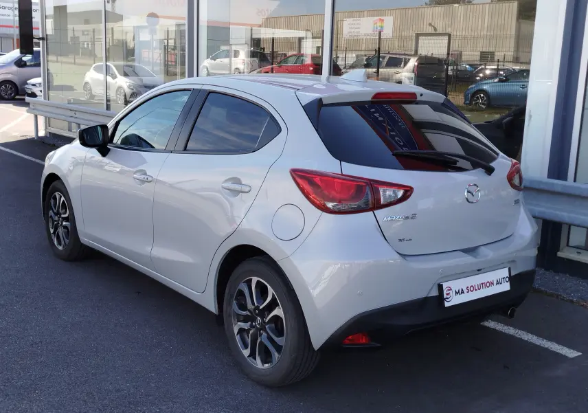 Vue 3/4 arrière droite d'une Mazda2 blanche nacrée avec jantes alliage et vitres teintées, stationnée devant un bâtiment vitré.