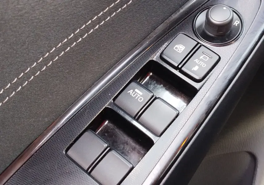 Gros plan sur les commandes de lève-vitres et réglage des rétroviseurs côté conducteur d'une Mazda2 blanche.