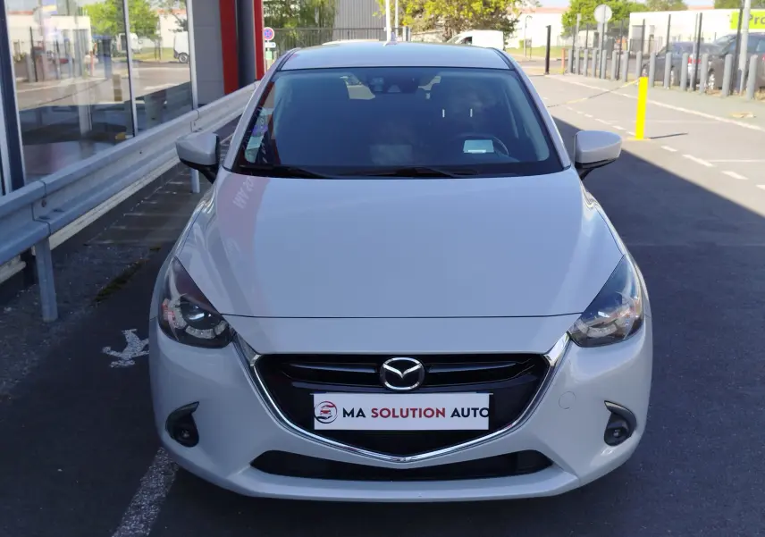 Vue avant d'une Mazda2 blanche nacrée garée devant un bâtiment, avec calandre chromée et phares allumés.
