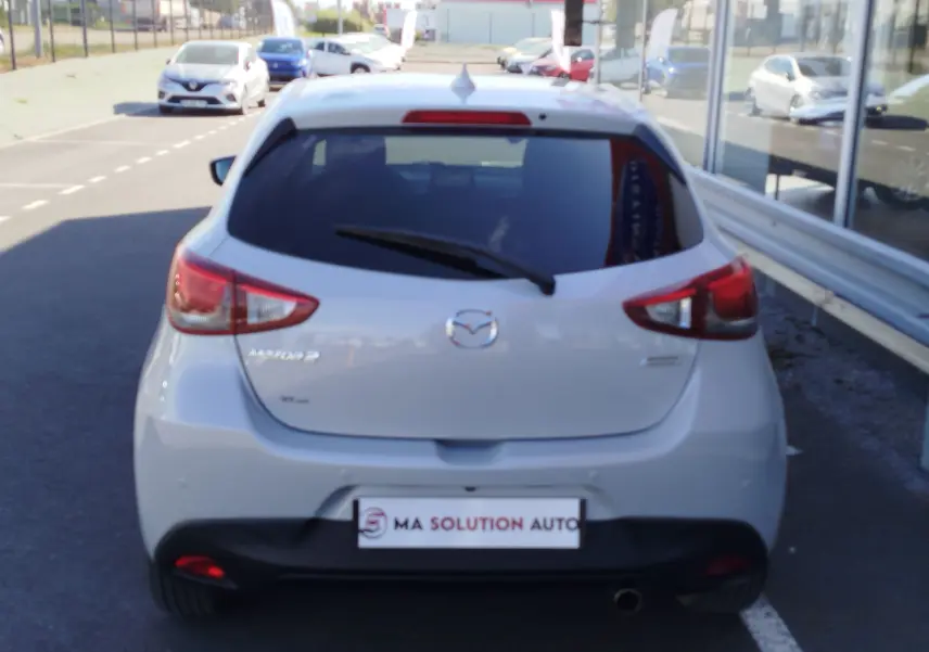 Vue arrière d'une Mazda2 blanche nacrée garée devant une vitrine avec logo et plaques visibles.