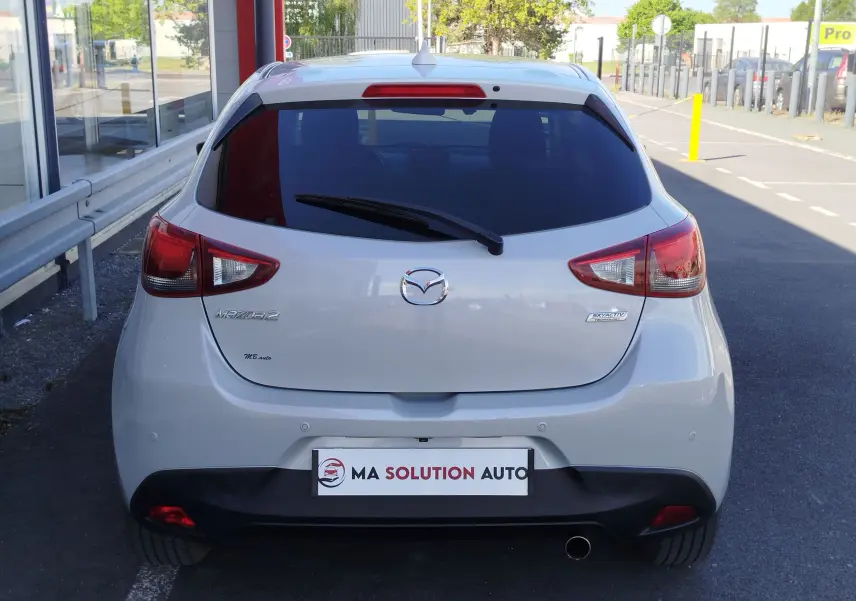 Vue arrière d'une Mazda2 blanche nacrée avec feux arrière rouges et logo Mazda bien visibles, stationnée en extérieur.