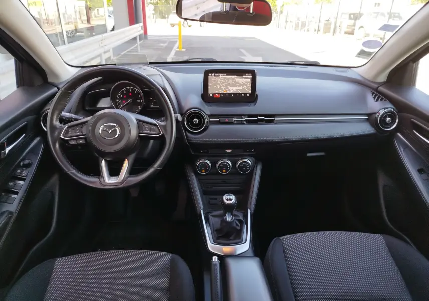 Vue intérieure avant de la Mazda2 2018 avec tableau de bord noir, écran central et boîte manuelle visible.