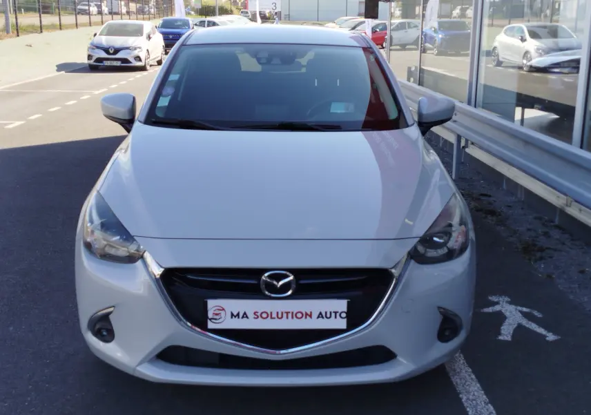 Vue avant d'une Mazda2 blanche nacrée stationnée devant un bâtiment avec logo Mazda visible sur la calandre.