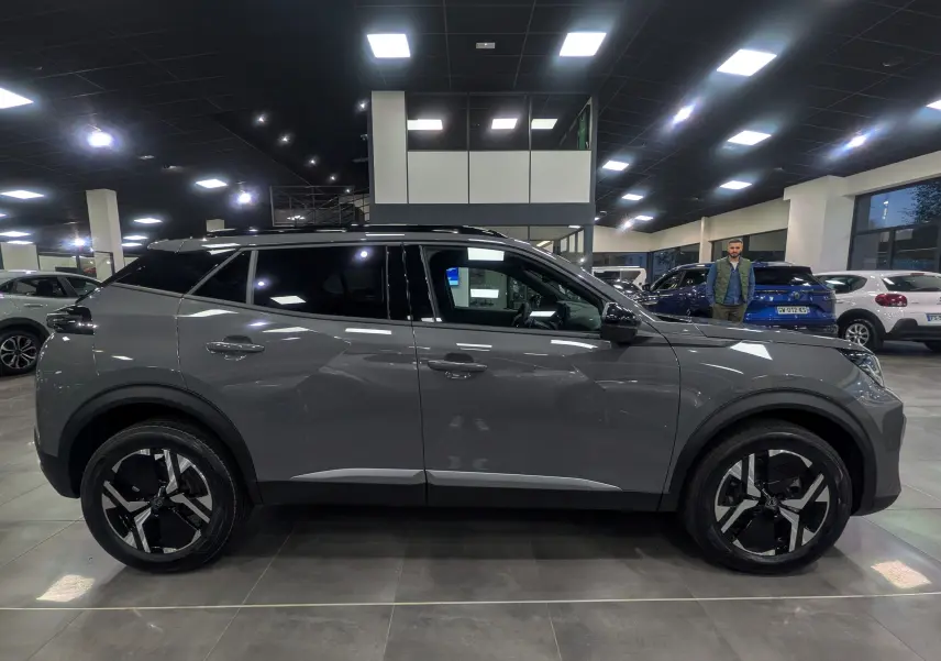 Profil côté droit du Peugeot 2008 Hybrid 145 gris Sélénium en showroom, soulignant ses jantes noires et toit noir contrastant.