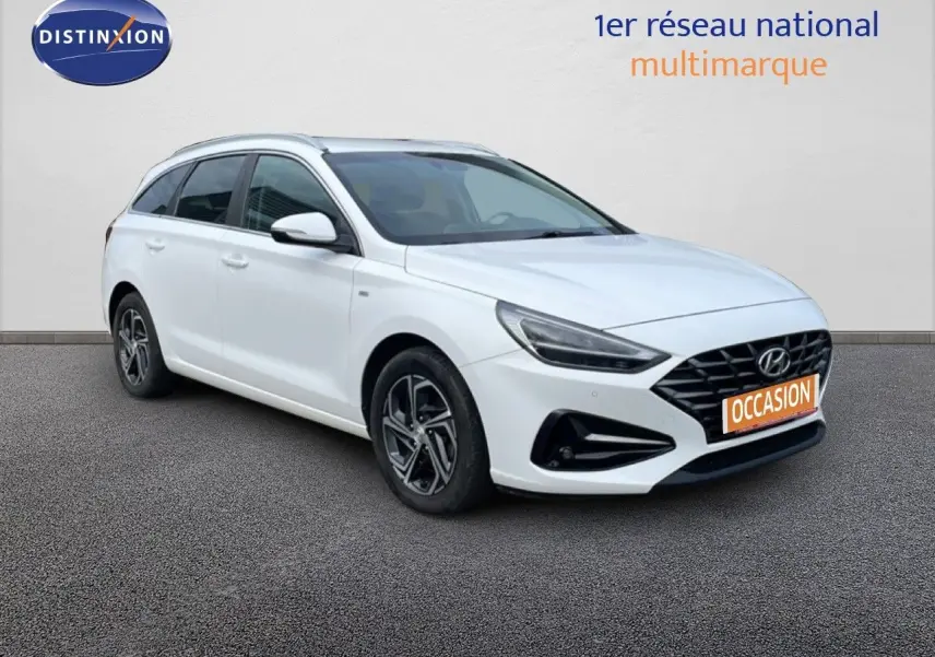 Vue 3/4 avant gauche d'une Hyundai i30 SW 2022 blanche Atlas avec calandre noire et jantes alliage distinctives.