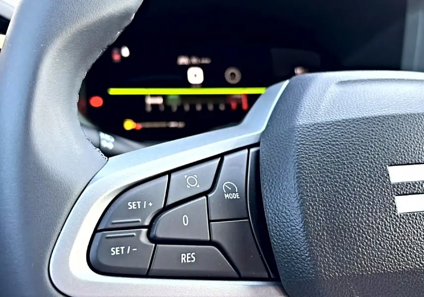 Gros plan sur les commandes multifonctions à gauche du volant noir du Dacia Bigster blanc, avec tableau de bord numérique flou en arrière-plan.