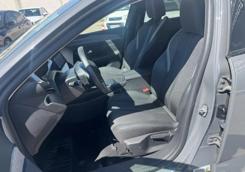 Intérieur avant droit de la Peugeot 308 Hybrid gris 2025, sièges tissu avec accoudoir central et tableau de bord moderne.