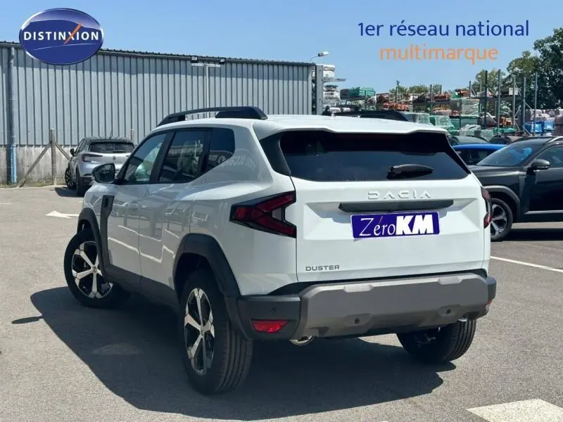 Vue 3/4 arrière droite d'un Dacia Duster blanc avec jantes alu et barres de toit, stationné en extérieur.