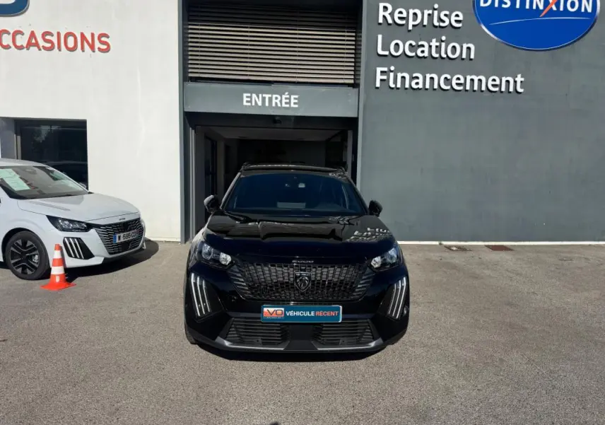 Vue avant d'un Peugeot 2008 Hybrid noir avec barres de toit, stationné devant une concession automobile.