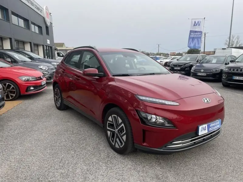 Hyundai Kona électrique rouge métal vu en 3/4 avant droit sur un parking de véhicules d'occasion.