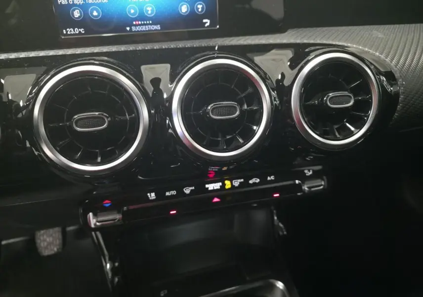 Gros plan sur les aérateurs ronds chromés et la console centrale noire du tableau de bord d'une Mercedes Classe A blanche.