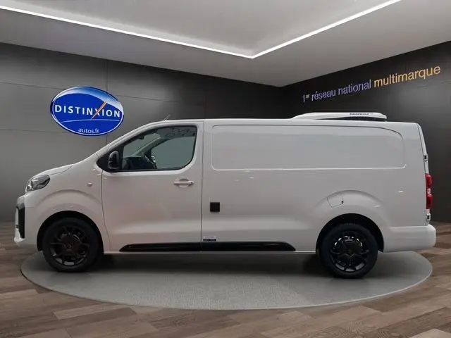 Vue latérale droite d'un utilitaire Citroën Jumpy blanc avec toit frigorifique et jantes noires en intérieur showroom.