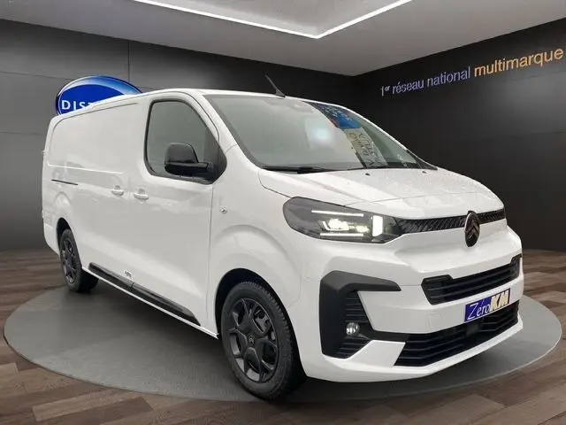 Vue 3/4 avant d’un Citroën Jumpy XL blanc avec phares allumés et jantes noires, en intérieur showroom.