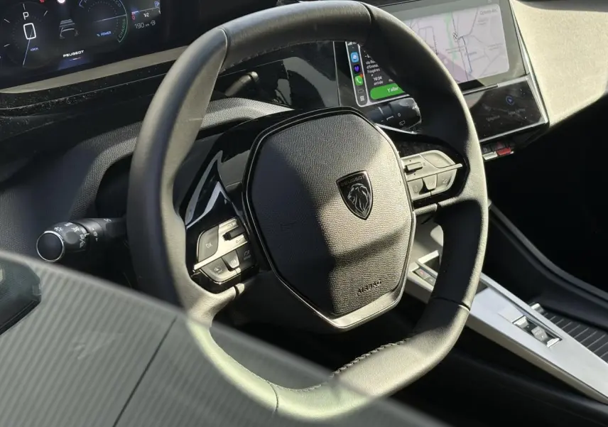 Vue rapprochée du volant cuir noir de la Peugeot 308 Hybrid 2025 avec tableau de bord numérique et écran tactile.