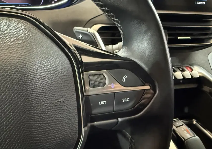 Gros plan sur le volant cuir noir du Peugeot 5008 2021, avec commandes audio et palette de changement de vitesse visible.
