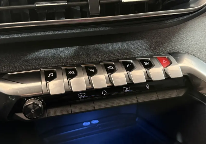 Gros plan sur la rangée de boutons argentés et noirs du tableau de bord du Peugeot 5008 gris clair, avec éclairage bleu dessous.