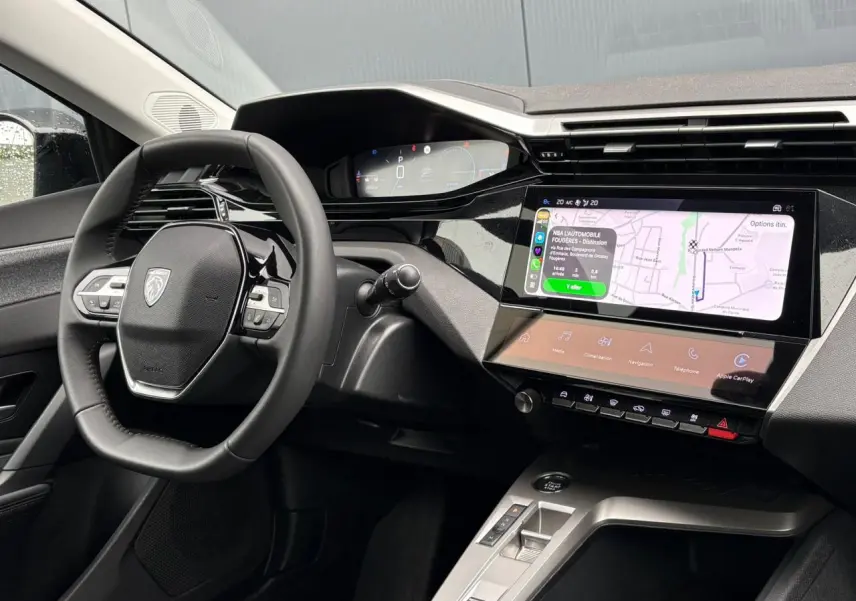 Vue intérieure du poste de conduite de la Peugeot 308 SW Hybrid 2025, avec écran tactile affichant la navigation et volant cuir noir.