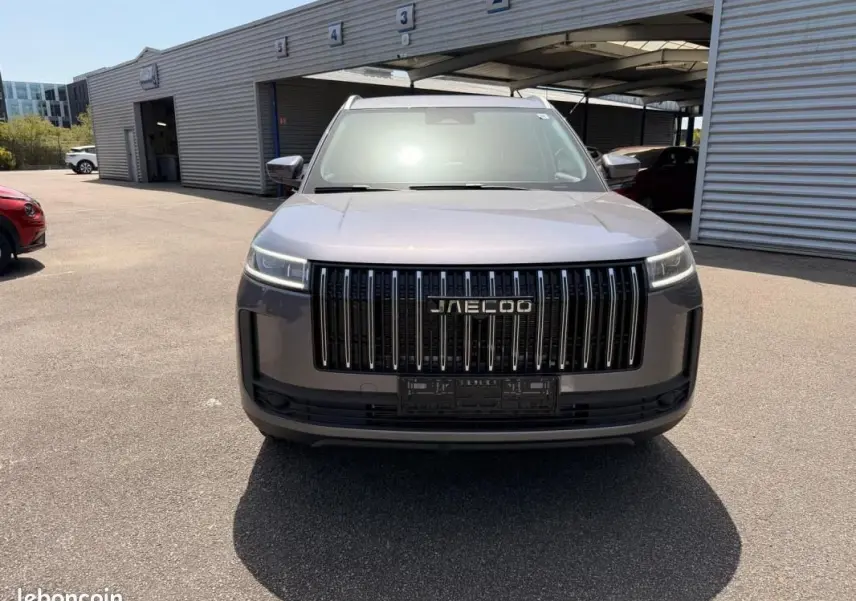 Vue 3/4 avant du JAECOO J5 gris Phantom avec calandre verticale noire et phares LED fins, stationné devant un bâtiment.