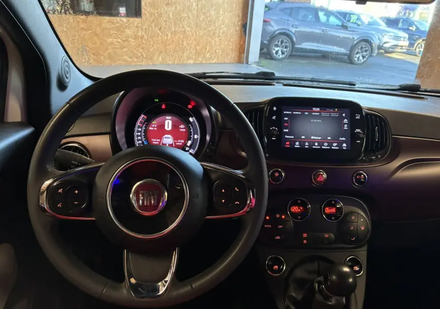 Vue intérieure centrée sur le volant et le tableau de bord de la Fiat 500 blanche, avec écran tactile et commandes visibles.