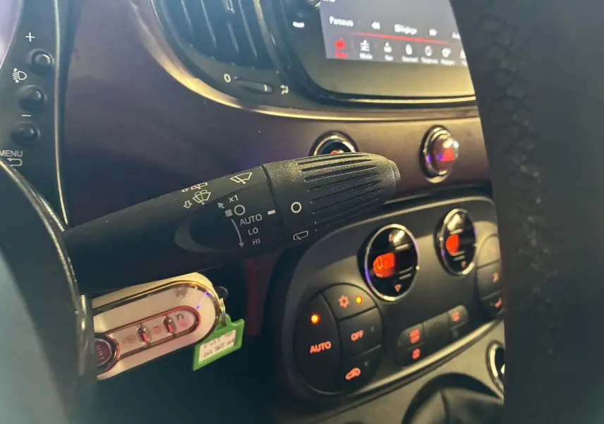 Gros plan intérieur sur la commande d’essuie-glace à gauche du volant d’une Fiat 500 blanche, avec tableau de bord et commandes clim visibles.