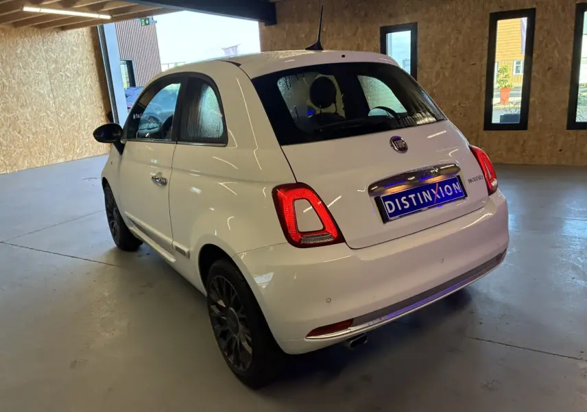 Vue 3/4 arrière droite d'une FIAT 500 blanche 2021, avec feux arrière allumés et jantes noires, stationnée en intérieur.