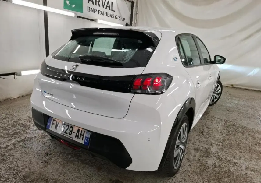 Vue 3/4 arrière droite d'une Peugeot 208 électrique blanche avec bandeau noir brillant entre feux arrière dans un garage.