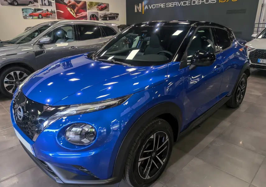 Nissan Juke Hybrid 2025 en bleu magnétique avec toit noir, vue 3/4 avant droit en intérieur showroom.