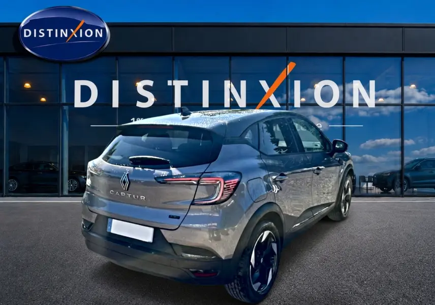 Vue 3/4 arrière d'un Renault Captur gris cassiopée 2026 avec jantes alu et feux arrière LED, devant une vitrine Distinxion.