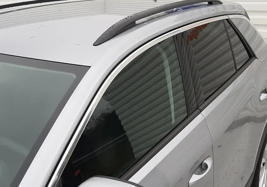Vue rapprochée du côté gauche avant du Volkswagen T-Roc gris pyrite métal, avec barres de toit noires et rétroviseur assorti.