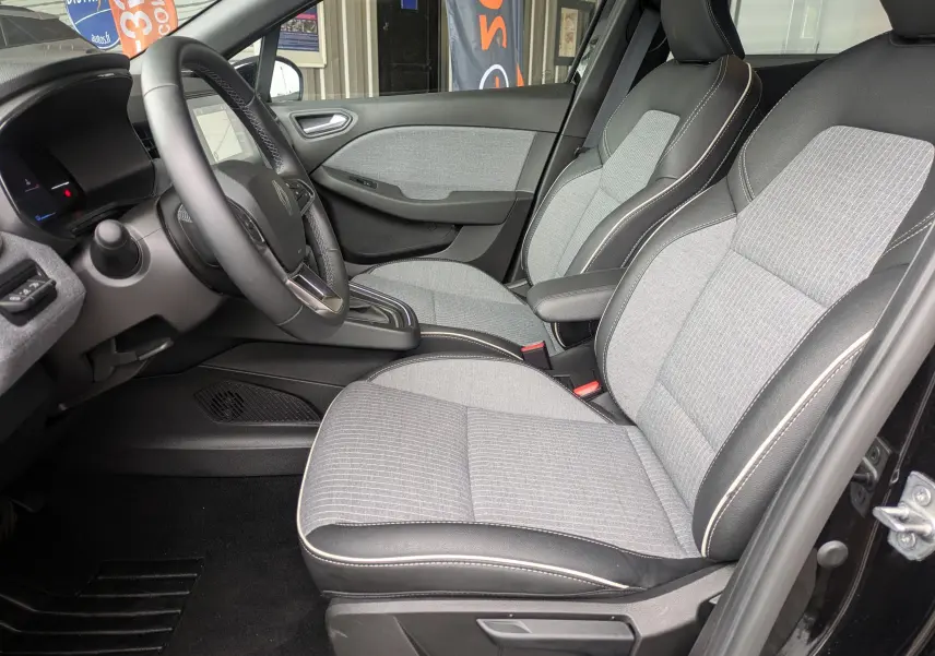 Vue intérieure côté gauche de la Renault Clio 2025, sièges tissu gris clair avec surpiqûres blanches et volant cuir noir.