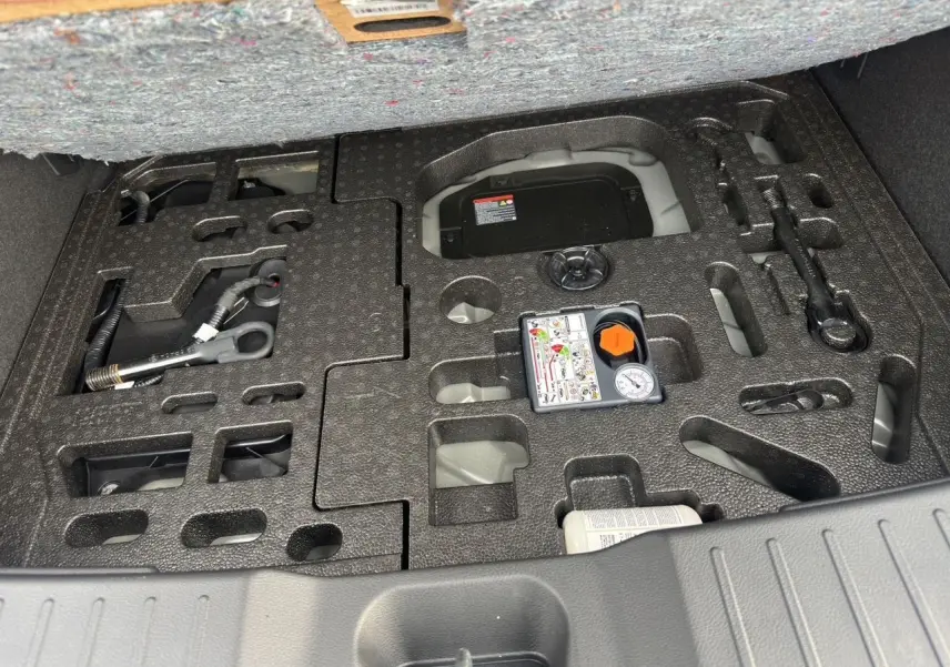 Coffre ouvert du Nissan Juke 2025 gris squale, vue plongeante sur le rangement d’outils et compresseur intégré.