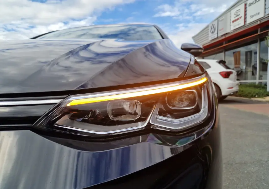 Gros plan sur le phare avant droit allumé d'une Volkswagen Golf 8 noire intense métal, avec reflet du ciel sur la carrosserie.