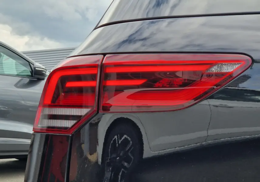 Gros plan sur le feu arrière droit d’une Volkswagen Golf 8 noire intense métal, reflétant un autre véhicule gris à côté.