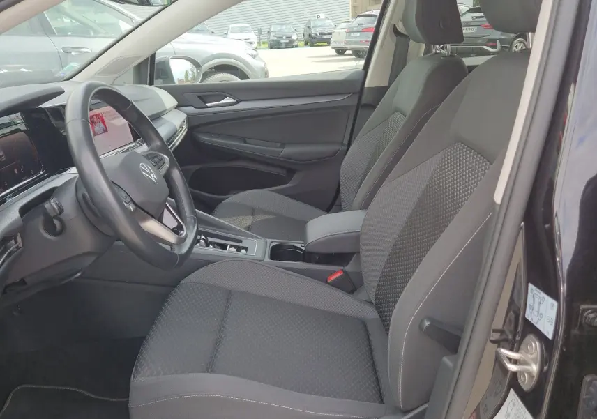 Vue intérieure côté conducteur de la Volkswagen Golf 8 noire, montrant les sièges avant et le tableau de bord moderne.