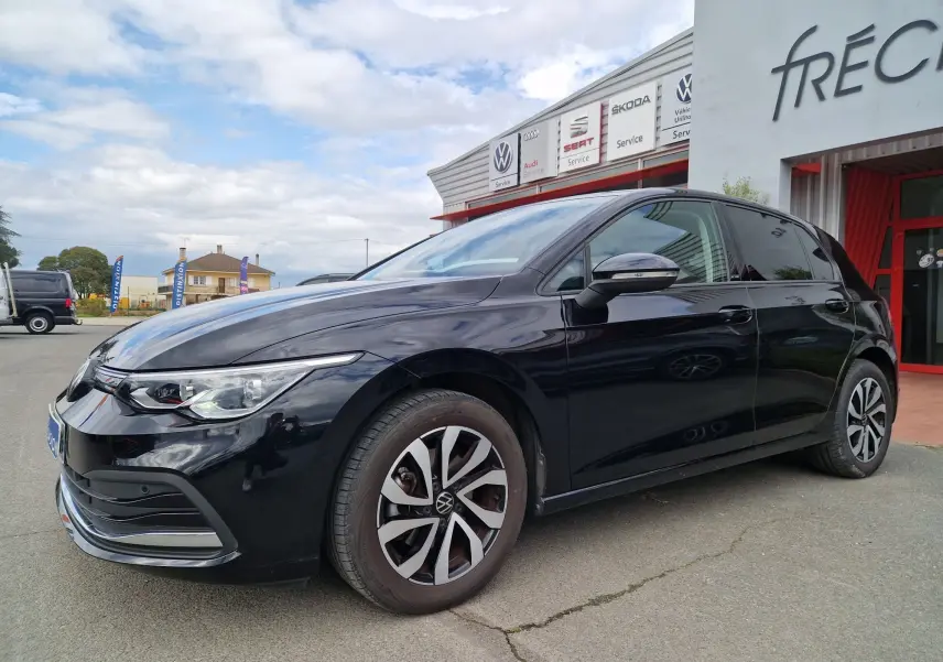 Vue 3/4 avant droit d'une Volkswagen Golf 8 noire intense métal garée devant un garage avec jantes distinctives.