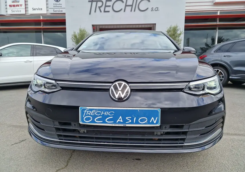 Vue frontale d'une Volkswagen Golf 8 noire intense métal, avec calandre et phares LED distinctifs, devant un concessionnaire.