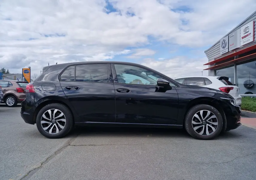 Vue latérale droite d'une Volkswagen Golf 8 noire intense métal, avec jantes alliage distinctives, stationnée devant un concessionnaire.
