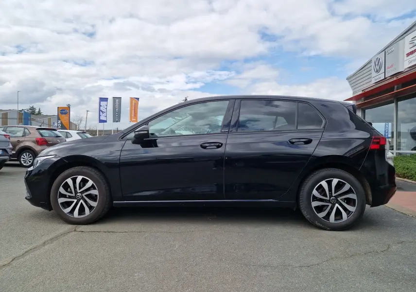 Profil côté gauche d'une Volkswagen Golf 8 noire intense métal, avec jantes distinctives et vitres teintées, stationnée en extérieur.