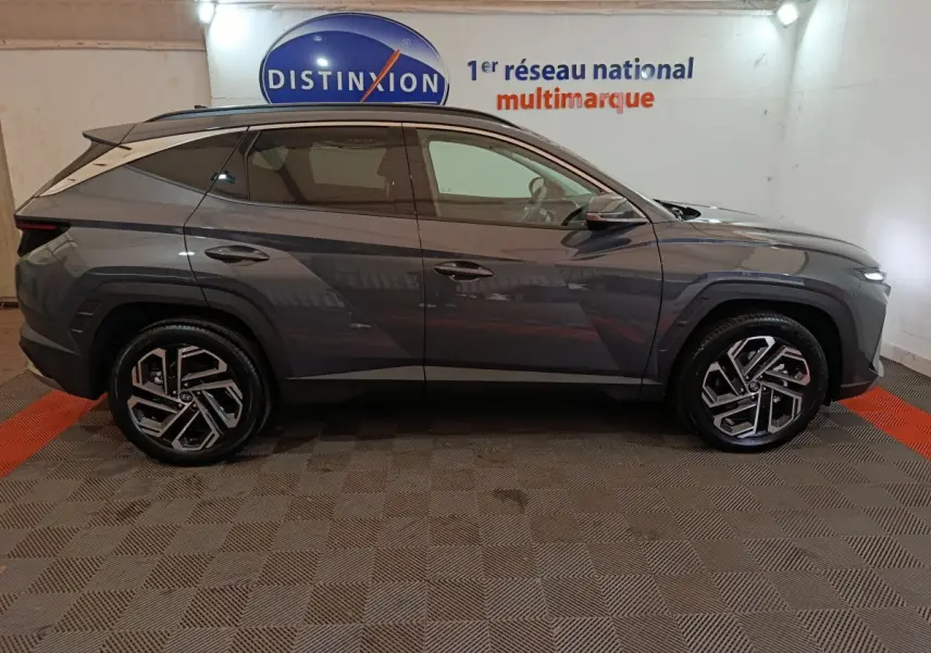 Profil côté gauche d'un Hyundai Tucson 1.6 Hybrid gris foncé 2025 avec jantes alliage et toit noir en intérieur showroom.
