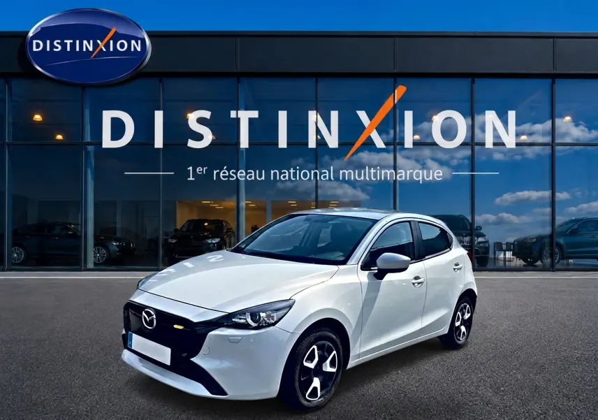 Vue 3/4 avant d'une Mazda2 blanche 2024 avec calandre noire et jantes distinctives, devant une vitrine Distinxion.