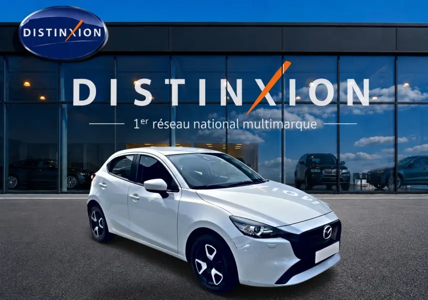 Vue 3/4 avant d'une Mazda2 blanche 2024 avec jantes noires et calandre noire devant un showroom Distinxion.