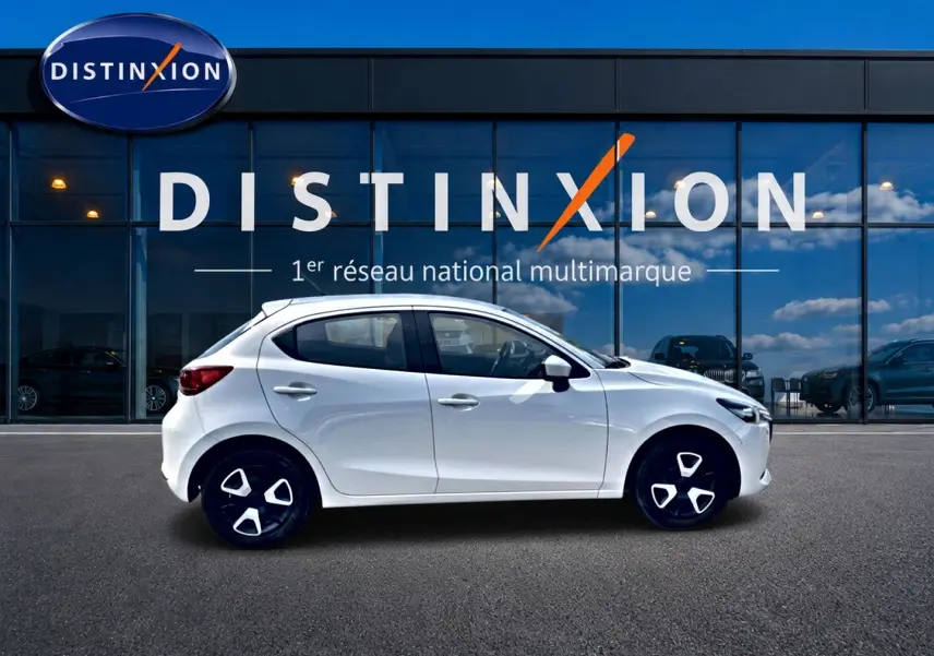 Vue latérale droite d'une Mazda2 blanche 2024 avec jantes noires et détails argentés, devant une vitrine Distinxion.