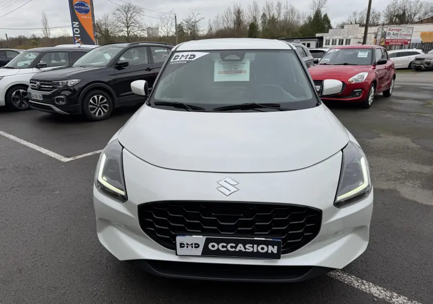 Vue avant d'une Suzuki Swift 1.2 Privilège Hybrid blanche avec calandre noire et phares LED allumés, en concession.