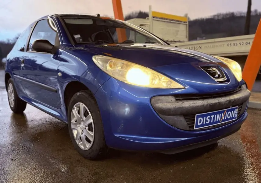 Peugeot 206+ bleu vue de profil côté gauche sous abri, phare avant allumé sur sol mouillé.