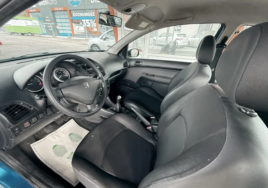 Peugeot 206+ bleu vue 3/4 avant droit avec phares allumés sur sol mouillé, devant structure orange.