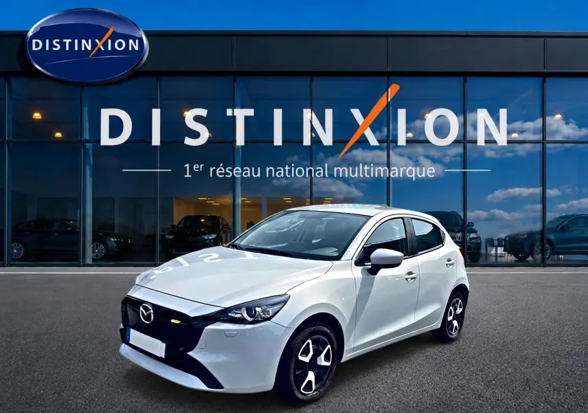 Vue 3/4 avant droite d'une Mazda2 blanche 2024 avec calandre noire et jantes au design distinctif, devant un showroom Distinxion.