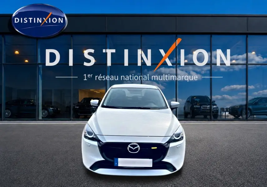 Vue frontale d'une Mazda2 blanche 2024 garée devant une vitrine avec logo Distinxion en arrière-plan.