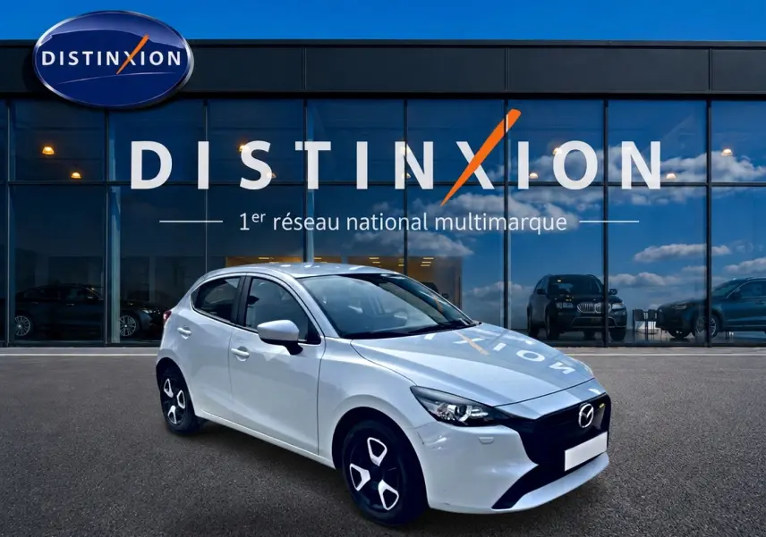 Vue 3/4 avant gauche d'une Mazda2 blanche 2024 avec calandre noire et jantes distinctives devant un showroom Distinxion.